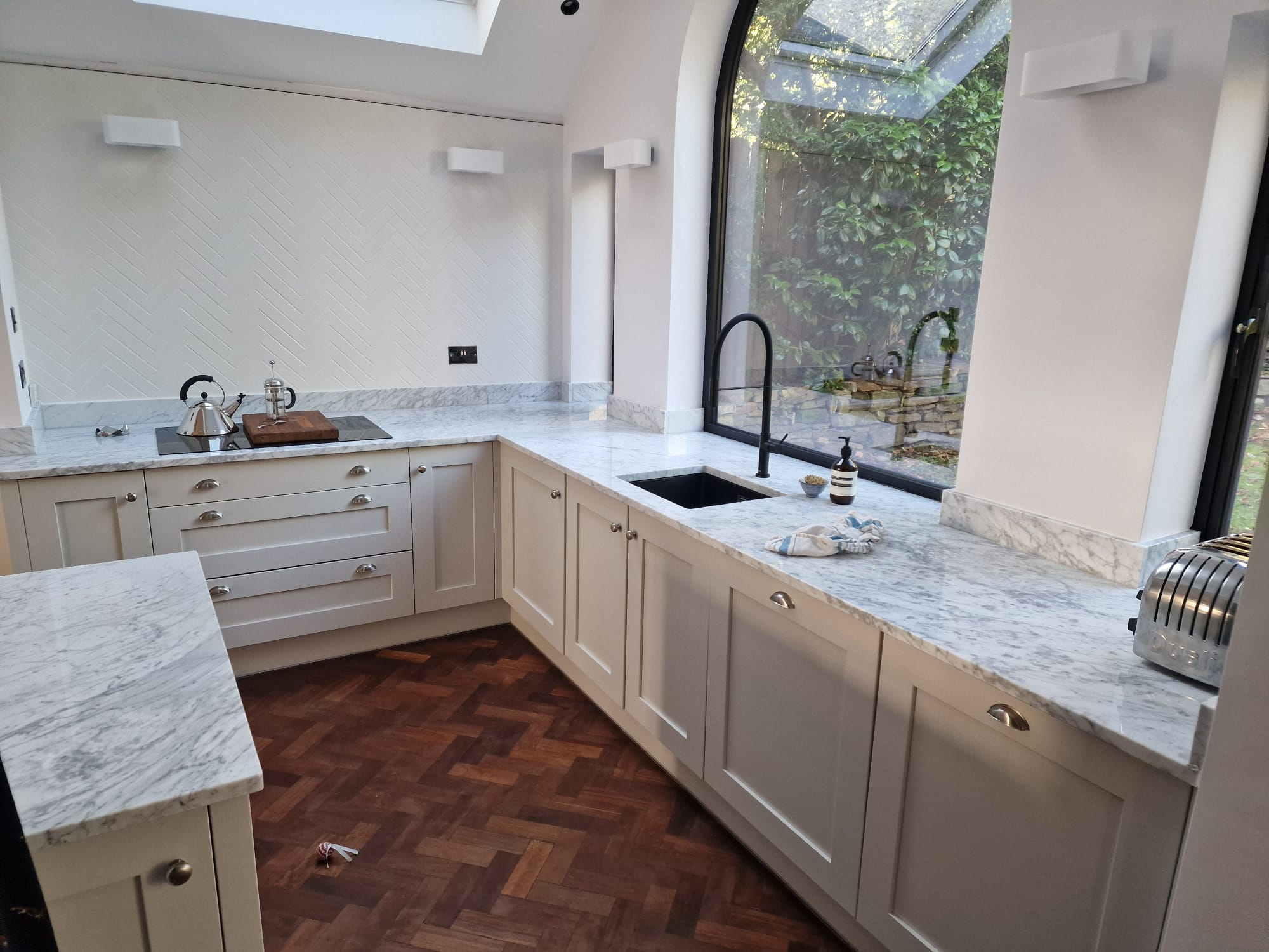 Premium fitted kitchen with cream shaker units, Carrara marble worktops, matt black tap, arched feature window and herringbone parquet floor